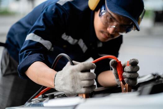 a man in a blue shirt and goggles is working on a car he is using a jumper cable to jump start the car photo